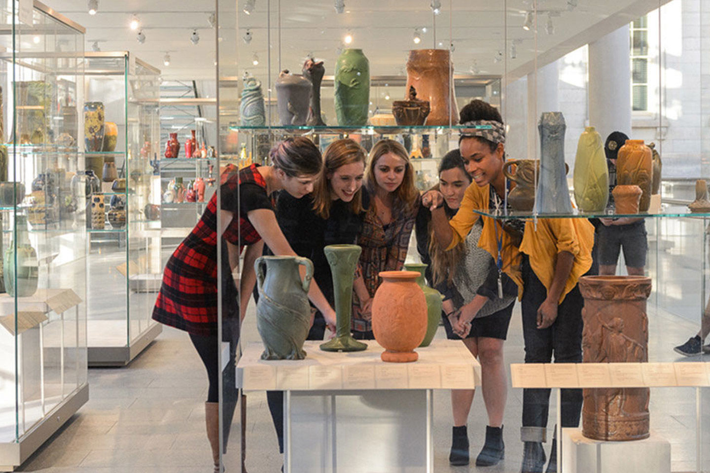 A group of teenagers are gathered round looking and pointing to a glass display case of vases within a light-filled gallery space